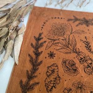 May include: A brown fabric tapestry with black ink illustrations of various plants, flowers, and other natural elements. The tapestry features a crescent moon and a circle of smaller moons, as well as stars.