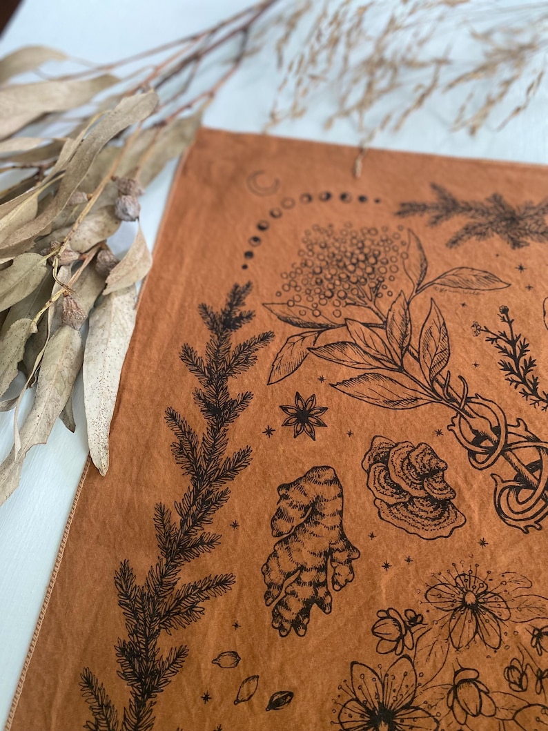 May include: A brown fabric tapestry with black ink illustrations of various plants, flowers, and other natural elements. The tapestry features a crescent moon and a circle of smaller moons, as well as stars.