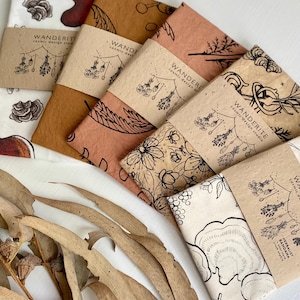 May include: Five different fabric bandanas with a nature-inspired design. Each bandana features a different botanical illustration, including mushrooms, flowers, and leaves. The bandanas are arranged in a stack, with the top bandana being a light brown color and the bottom bandana being a white color.