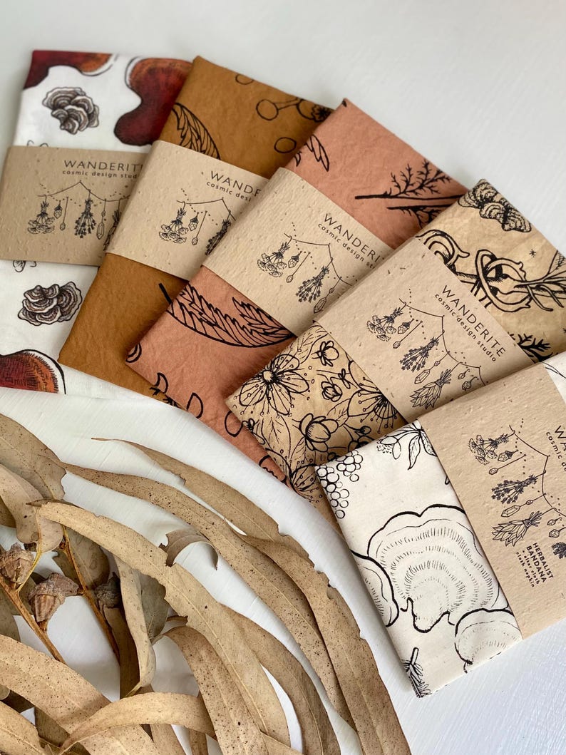 May include: Five different fabric bandanas with a nature-inspired design. Each bandana features a different botanical illustration, including mushrooms, flowers, and leaves. The bandanas are arranged in a stack, with the top bandana being a light brown color and the bottom bandana being a white color.