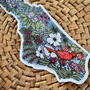 May include: A sticker in the shape of a state, featuring a red cardinal bird perched among various flowers and greenery. The design includes white, pink, and purple flowers, with a light blue background. The sticker is placed on a woven surface.