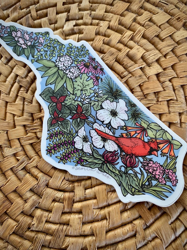 May include: A sticker in the shape of a state, featuring a red cardinal bird perched among various flowers and greenery. The design includes white, pink, and purple flowers, with a light blue background. The sticker is placed on a woven surface.
