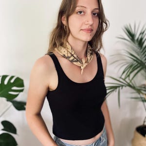 May include: A woman wearing a black tank top with a light brown bandana tied around her neck. She is wearing blue jeans and has long brown hair.