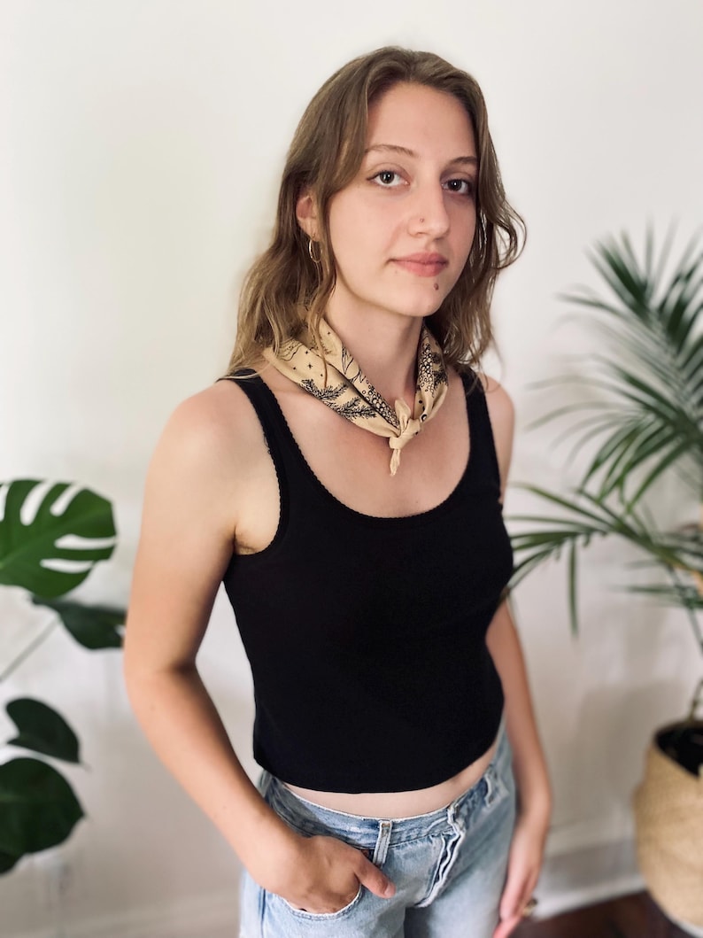 May include: A woman wearing a black tank top with a light brown bandana tied around her neck. She is wearing blue jeans and has long brown hair.