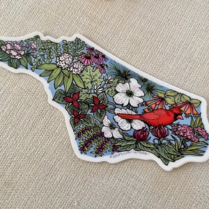 May include: A sticker in the shape of a state, filled with colorful flowers, green leaves, and a red cardinal. The background is light blue, and the sticker has a white border. The sticker is on a neutral-colored surface.