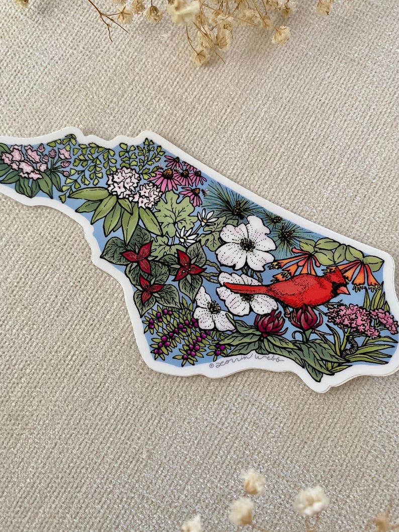 May include: A sticker in the shape of a state, filled with colorful flowers, green leaves, and a red cardinal. The background is light blue, and the sticker has a white border. The sticker is on a neutral-colored surface.