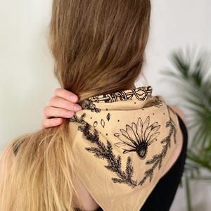 May include: A beige bandana with a black floral design featuring a daisy and pine branches. The bandana is tied around the neck of a person with long blonde hair.