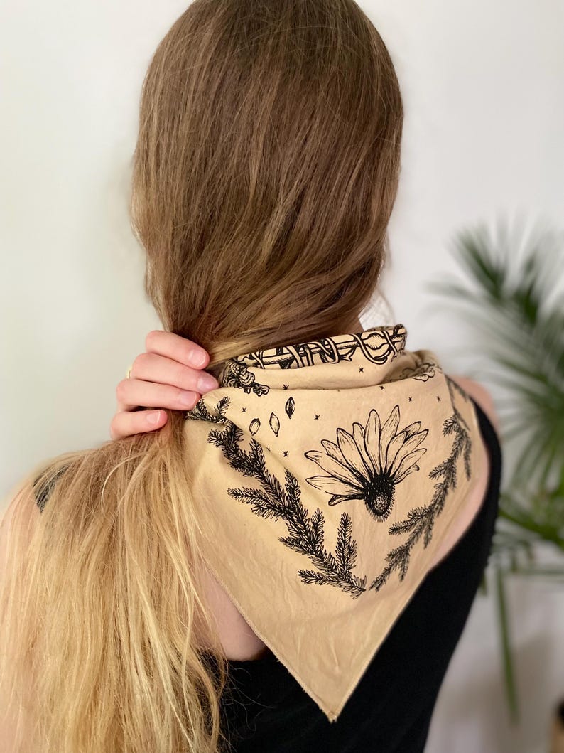 May include: A beige bandana with a black floral design featuring a daisy and pine branches. The bandana is tied around the neck of a person with long blonde hair.