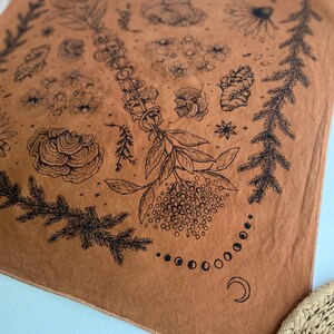 May include: A brown fabric bandana with a black and white design of various plants, flowers, and a crescent moon. The design is centered in a circle with a border of pine branches.