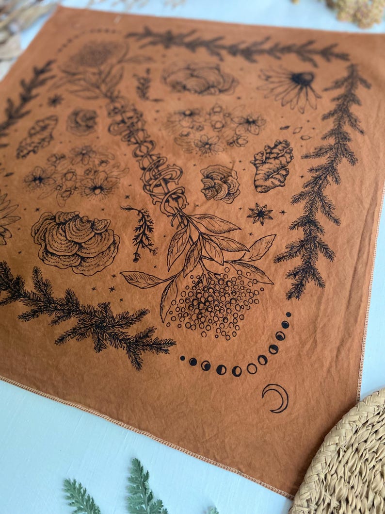 May include: A brown fabric bandana with a black and white design of various plants, flowers, and a crescent moon. The design is centered in a circle with a border of pine branches.