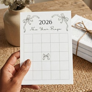 Puede incluir: Una tarjeta de bingo de A&ntilde;o Nuevo 2026 blanca con una cuadr&iacute;cula, sostenida por una mano. La tarjeta presenta ilustraciones de lazos y texto. Un jarr&oacute;n con vegetaci&oacute;n y una pila de libros atados con hilo est&aacute;n en el fondo.