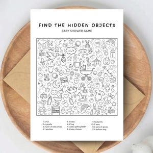 Peut inclure: Une carte de jeu de baby shower en noir et blanc avec le texte "FIND THE HIDDEN OBJECTS BABY SHOWER GAME". La carte présente une illustration détaillée d'articles pour bébés. La carte est sur une assiette en bois avec une enveloppe marron.