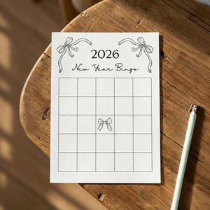 Puede incluir: Una tarjeta de bingo de A&ntilde;o Nuevo blanca con una cuadr&iacute;cula de cuadrados, con el a&ntilde;o 2026 y el texto "New Year Bingo". Ilustraciones de lazos decorativos en la parte superior y central. Un l&aacute;piz verde claro descansa sobre la superficie de madera.