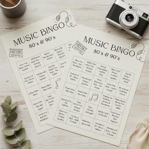 May include: Two Music Bingo cards with 80s & 90s music themes. The cards have a beige background with black text and illustrations of a cassette tape and headphones. A vintage camera and a small wooden pencil holder are also visible.