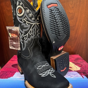 Könnte beinhalten: Schwarze Cowboystiefel aus Leder mit quadratischer Zehenpartie und weißen Ziernähten am Schaft und Vorderblatt. Die Stiefel haben ein gelbes Innenfutter, einen gestapelten Holzabsatz und eine schwarze Gummilaufsohle mit roten Akzenten. Ein La Sierra Markenetikett ist befestigt.
