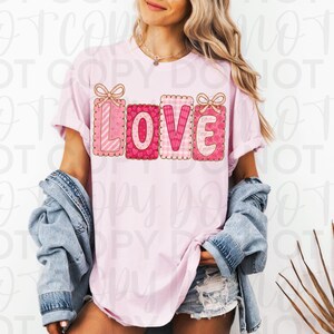 May include: A light pink t-shirt with the word "LOVE" in a decorative, gift-box style design. The letters are in shades of pink and red with bow accents. The shirt is worn with a denim jacket and shorts.