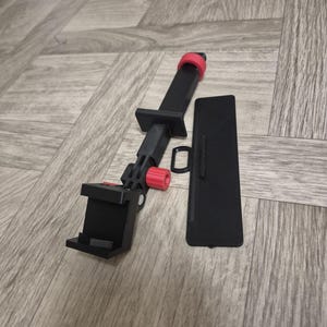 以下が含まれることがあります： 黒と赤の調節可能な電話ホルダーと、別の黒い長方形の部品。 ホルダーには電話を固定するためのクランプと赤い調整ノブが付いています。 木の表面に置かれたアイテムです。