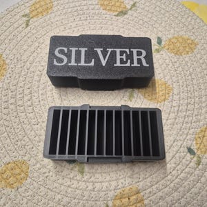 May include: A black rectangular box with the word "SILVER" in white lettering is displayed above a grey rectangular container with vertical dividers. The items are on a beige placemat with a pineapple pattern.