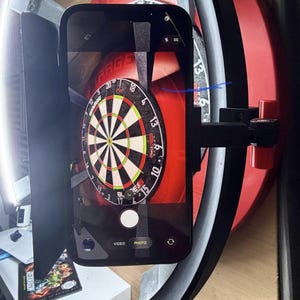 May include: A smartphone is mounted in front of a dartboard, capturing a photo. The dartboard is red and black with white and green accents. The phone is black and has a camera app open. A black bracket holds the phone in place.