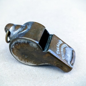 May include: A vintage brass whistle with a tarnished finish. The whistle has a ring for attachment and the text "The Acme Thunderer" is visible. The overall color is a mix of brown and silver.