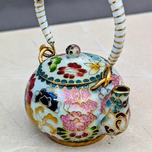 May include: A miniature teapot with a white handle and a floral pattern. The teapot has a light blue base with pink, red, yellow, and blue flowers. Gold detailing is used throughout the design, adding to its decorative appeal.