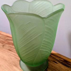 May include: A light green glass vase in a tulip shape. The vase features a textured leaf pattern and rests on a round base. The vase is displayed on a wooden surface.