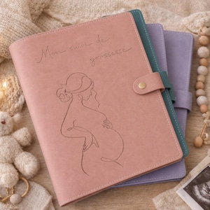 Peut inclure: Un carnet rose poudré avec une fermeture à pression dorée, orné d'un dessin au trait représentant une silhouette enceinte et le texte Mon suivi de grossesse. Deux autres carnets, bleu canard et lilas, sont placés derrière avec leurs fermetures.