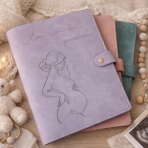 Peut inclure: Un journal lavande avec le texte "Mon suivi de grossesse" et une illustration en relief d'une personne enceinte. Le journal a une fermeture à bouton-pression et est à côté de journaux roses et verts.