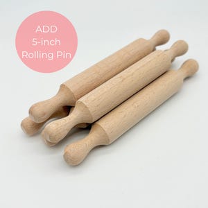 May include: Four wooden rolling pins, each approximately 5 inches long, are arranged on a white surface. The rolling pins are made of light-colored wood and have a classic design with rounded handles. A pink circle with the text "ADD 5-inch Rolling Pin" is in the upper left corner.