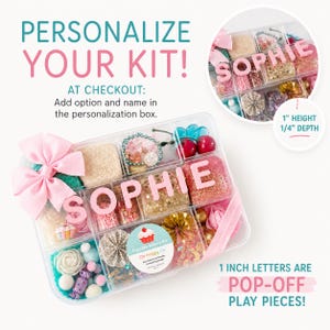 May include: A clear plastic organizer box filled with various craft supplies, including pink letters spelling SOPHIE, a pink bow, red cherry charms, gold paper fans, and a sticker labeled Cupcake Bakery Kit Oh Happy Co. The letters are 1 inch high.