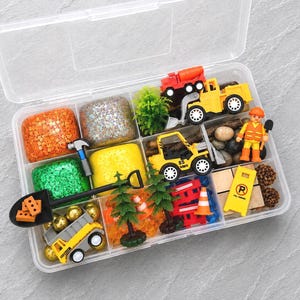 May include: A clear plastic storage box filled with construction-themed toys and sensory materials. Includes toy trucks, a construction worker figure, play dough, small trees, rocks, and a road sign. The play set features bright colors and various textures.