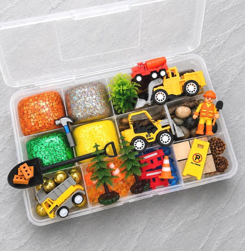May include: A clear plastic storage box filled with construction-themed toys and sensory materials. Includes toy trucks, a construction worker figure, play dough, small trees, rocks, and a road sign. The play set features bright colors and various textures.