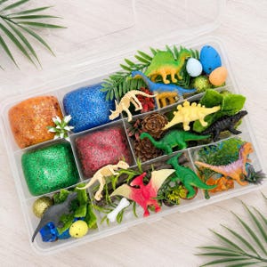May include: A clear plastic storage container filled with various sensory play items including four blocks of glittery slime in orange, blue, green, and red, multiple plastic dinosaur figurines, small artificial plants, pinecones, and colorful eggs.