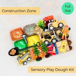 May include: A sensory play dough kit labeled "Construction Zone" with various construction-themed toys and colorful play dough. Includes toy trucks, a construction worker figure, and miniature trees. The kit is in a clear plastic container.