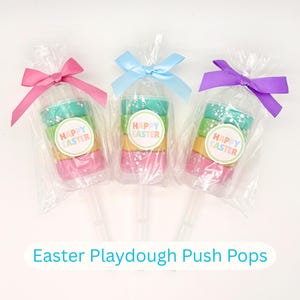 May include: Three Easter playdough push pops in clear plastic packaging, each topped with a colorful bow. The playdough is layered in green, yellow, and pink, with a "Happy Easter" sticker. The text "Easter Playdough Push Pops" is at the bottom.