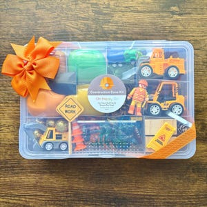 May include: A clear plastic container filled with construction-themed toys and sensory play dough. The kit includes toy trucks, a road sign, cones, and a worker figurine. An orange bow adorns the container. The text on the container reads "Construction Zone Kit".