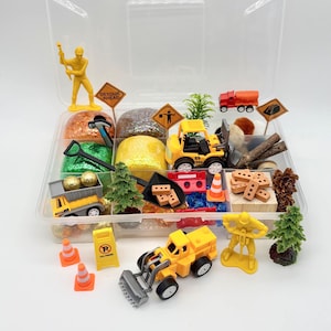 May include: A clear plastic container filled with miniature construction-themed toys and accessories. Includes yellow construction vehicles, traffic cones, signs, trees, and glittery play dough. The set is designed for imaginative play and creative exploration.