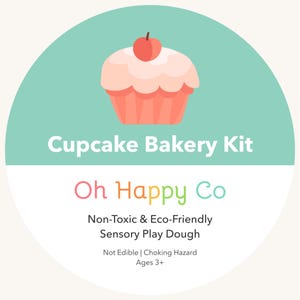May include: A circular label for a Cupcake Bakery Kit by Oh Happy Co. The top half is mint green with a pink cupcake illustration featuring a red cherry. The bottom half is white with text: Non-Toxic & Eco-Friendly Sensory Play Dough, Not Edible, Choking Hazard, Ages 3+.