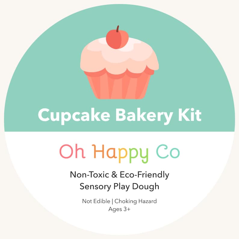 May include: A circular label for a Cupcake Bakery Kit by Oh Happy Co. The top half is mint green with a pink cupcake illustration featuring a red cherry. The bottom half is white with text: Non-Toxic & Eco-Friendly Sensory Play Dough, Not Edible, Choking Hazard, Ages 3+.