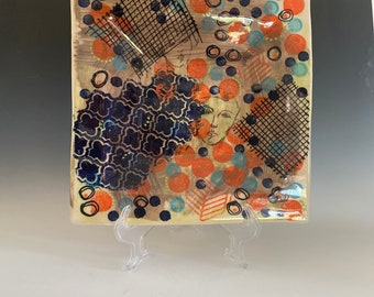 May include: A square ceramic plate with a colorful abstract design. The design features orange, blue, and black circles, as well as a black and white grid pattern.