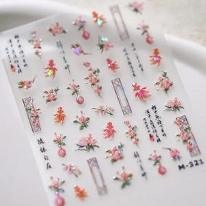 May include: A sheet of adhesive nail decals featuring iridescent pink floral motifs, orange goldfish, rectangular geometric frames, and black Chinese calligraphy characters. The stickers have a glossy, reflective finish and are arranged in a grid pattern on a clear backing sheet labeled M-321.
