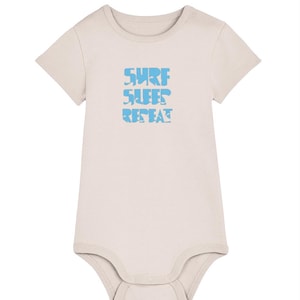 May include: A light beige short-sleeved baby bodysuit featuring a crew neckline and a snap-button closure at the bottom. The center of the chest displays the text "SURF SLEEP REPEAT" printed in a light blue color with a distressed, wavy texture.