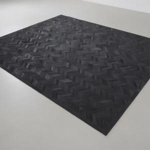 Puede incluir: Una alfombra negra rectangular grande con un patrón de espiga. La superficie de la alfombra está formada por pequeños bloques rectangulares dispuestos en un diseño en forma de V repetida. La alfombra está colocada sobre una superficie blanca.