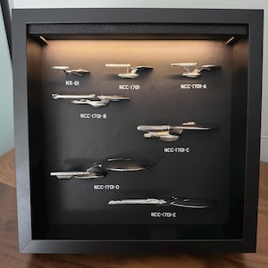 K&ouml;nnte beinhalten: Eine schwarze, gerahmte Vitrine mit sieben silbernen Modellraumschiffen. Jedes Schiff ist mit Text beschriftet, darunter "NX-01" und "NCC-1701" gefolgt von Buchstaben. Die Schiffe sind vor einem schwarzen Hintergrund mit warmem Licht platziert.