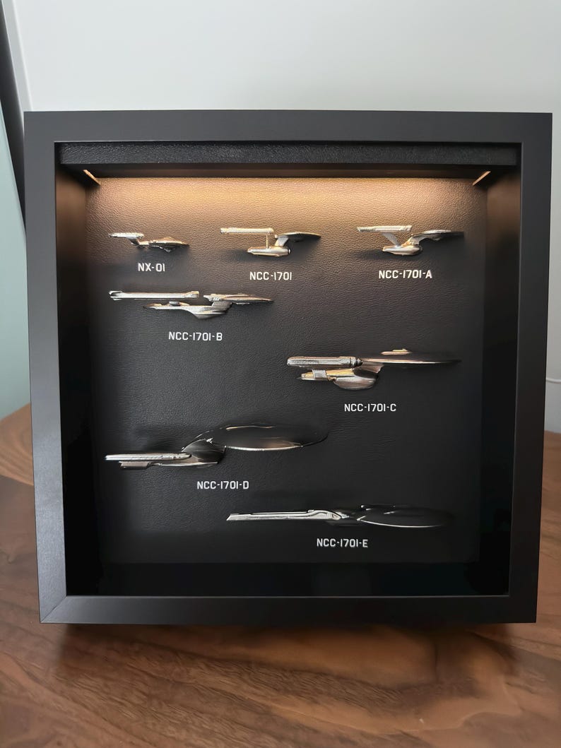 K&ouml;nnte beinhalten: Eine schwarze, gerahmte Vitrine mit sieben silbernen Modellraumschiffen. Jedes Schiff ist mit Text beschriftet, darunter "NX-01" und "NCC-1701" gefolgt von Buchstaben. Die Schiffe sind vor einem schwarzen Hintergrund mit warmem Licht platziert.