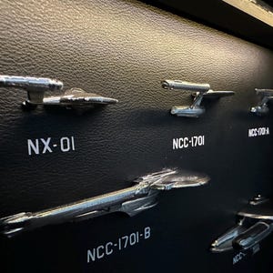 K&ouml;nnte beinhalten: Eine Sammlung silberfarbener Modellraumschiffe, darunter NX-01 und NCC-1701, montiert auf einer schwarzen, strukturierten Oberfl&auml;che. Die Modelle sind detaillierte Nachbildungen von Raumschiffen aus einer Science-Fiction-Serie.
