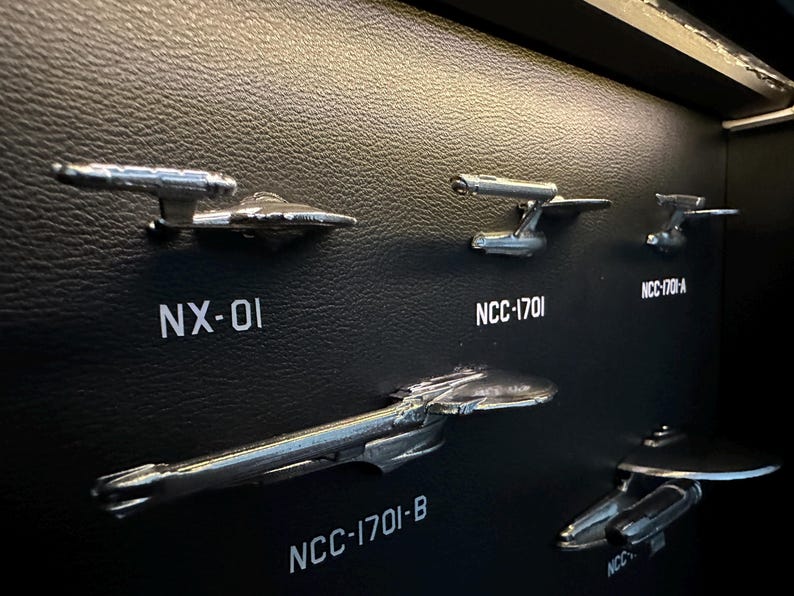 K&ouml;nnte beinhalten: Eine Sammlung silberfarbener Modellraumschiffe, darunter NX-01 und NCC-1701, montiert auf einer schwarzen, strukturierten Oberfl&auml;che. Die Modelle sind detaillierte Nachbildungen von Raumschiffen aus einer Science-Fiction-Serie.