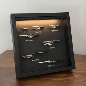 K&ouml;nnte beinhalten: Ein gerahmtes Display mit Miniatur-Metallraumschiffen, darunter NX-01, NCC-1701 und die Varianten A, B, C, D und E. Die Schiffe sind auf einem schwarzen Hintergrund in einem schwarzen Rahmen montiert und werden von einem warmen Licht beleuchtet.
