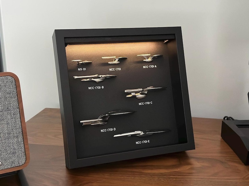 K&ouml;nnte beinhalten: Ein gerahmtes Display mit Miniatur-Metallraumschiffen, darunter NX-01, NCC-1701 und die Varianten A, B, C, D und E. Die Schiffe sind auf einem schwarzen Hintergrund in einem schwarzen Rahmen montiert und werden von einem warmen Licht beleuchtet.