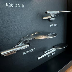 K&ouml;nnte beinhalten: Eine Pr&auml;sentation von vier silberfarbenen Raumschiffmodellen, die jeweils mit "NCC-1701" und einem Buchstaben gekennzeichnet sind. Die Modelle sind auf einer strukturierten schwarzen Oberfl&auml;che montiert, mit wei&szlig;em Text. Die Schiffe befinden sich in einer schwarzen Vitrine.